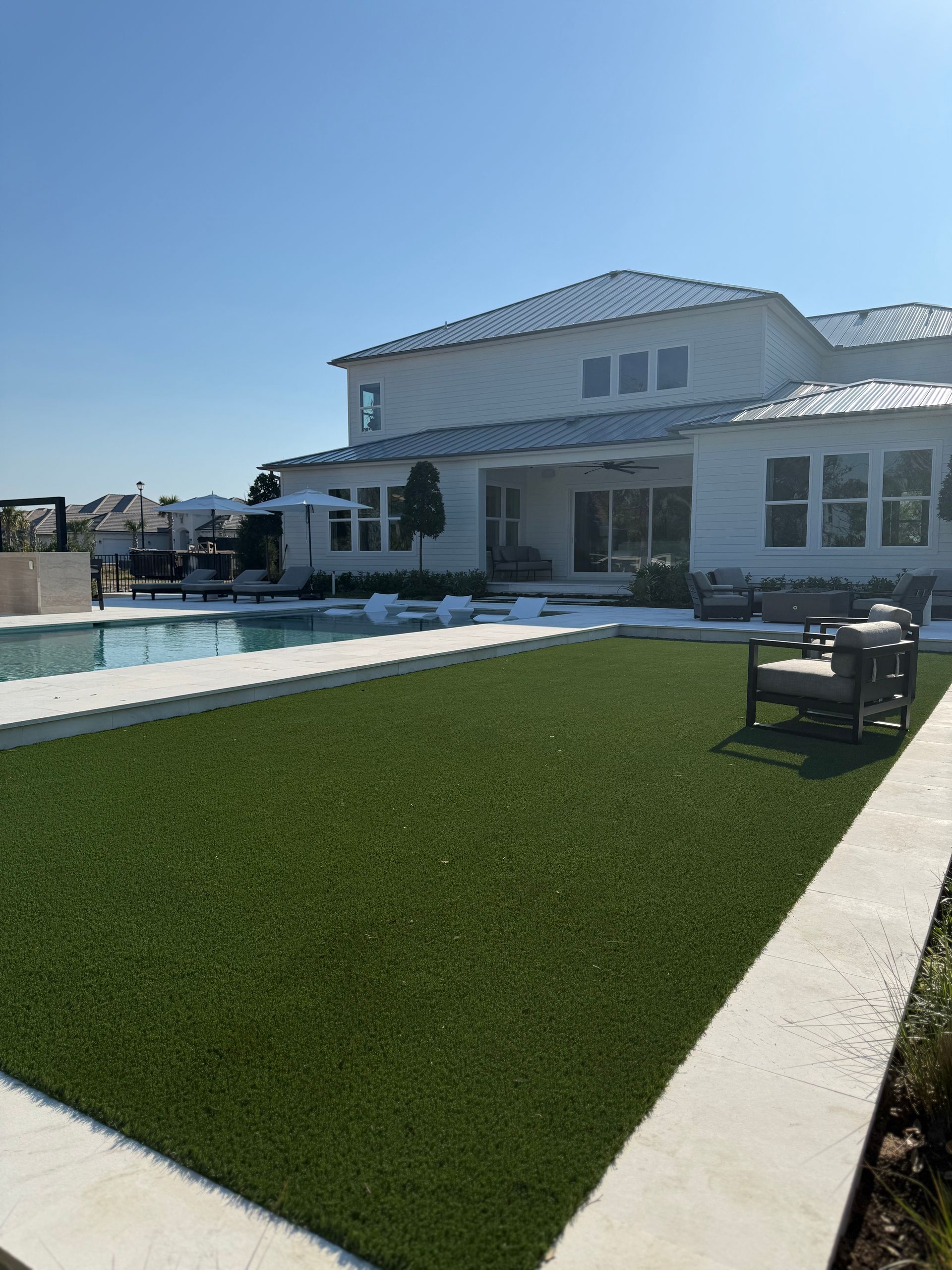 A spacious backyard with green turf, a pool, and a two-story white house under a clear blue sky.
