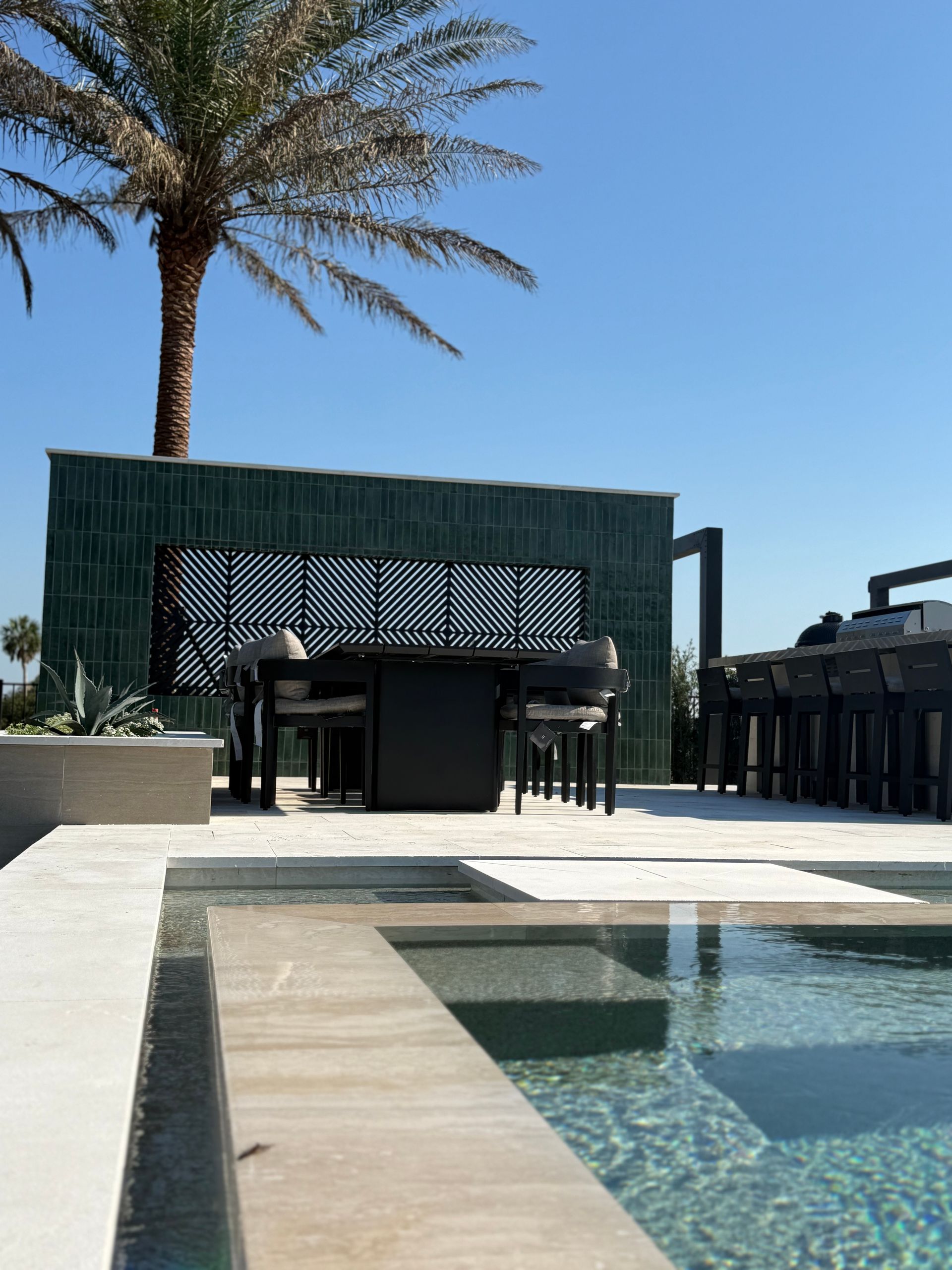 Outdoor dining area with a pool; palm tree, green wall, table, chairs, and blue sky.