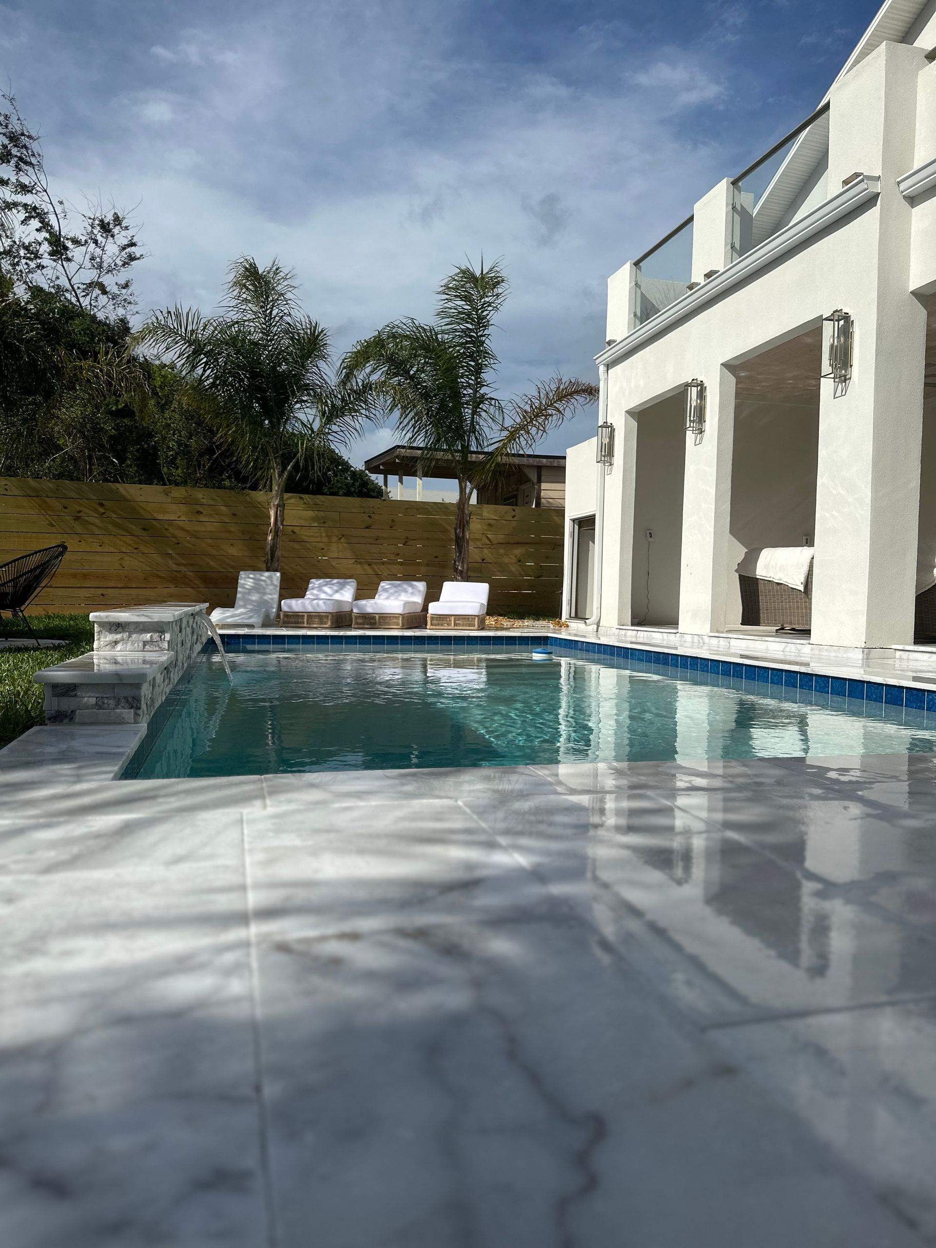 Pool area with white house, pool, chairs, trees, and cloudy sky.