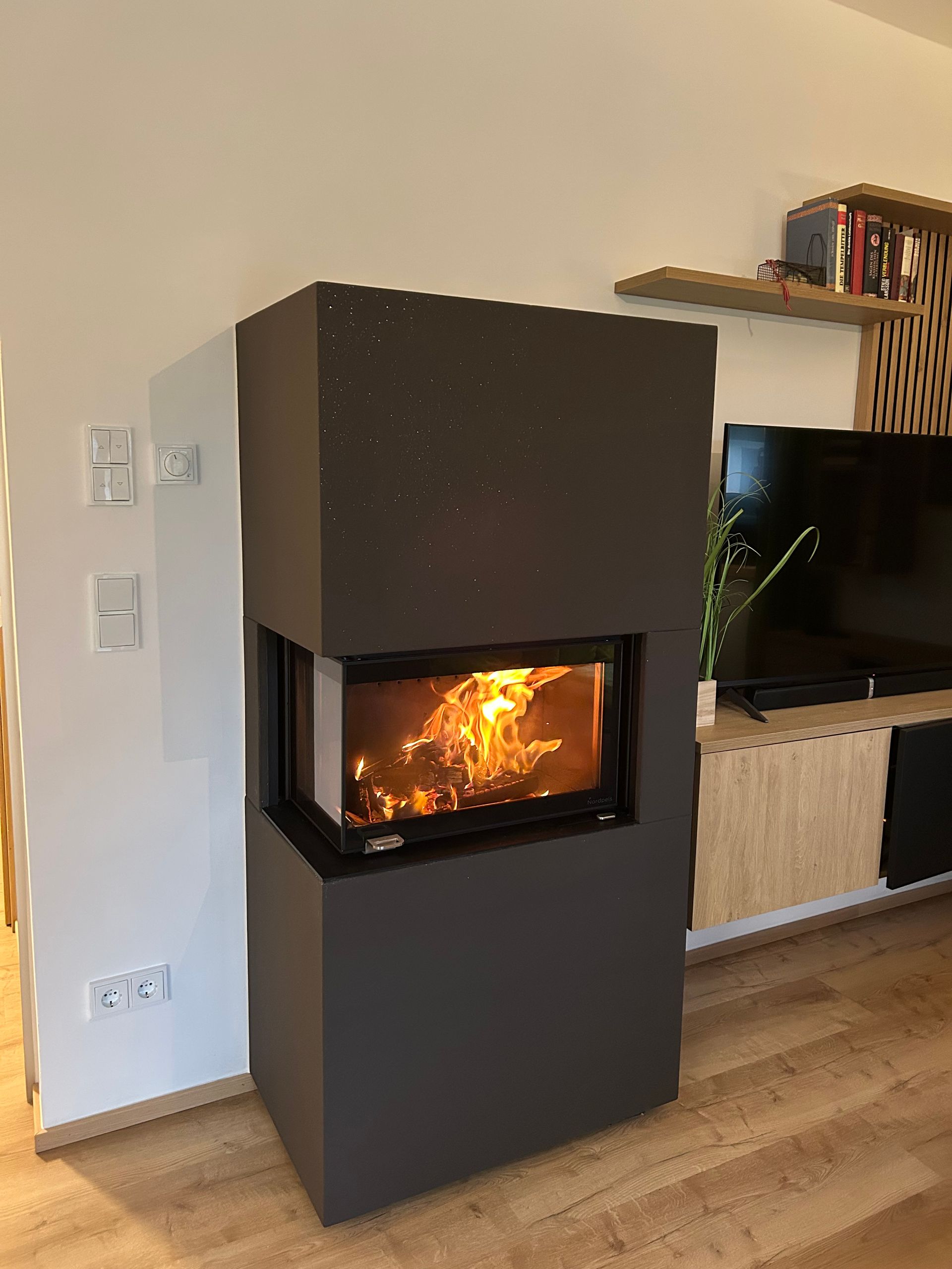 Moderner schwarzer Kamin mit brennenden Flammen im Wohnzimmer, neben einem Fernseher auf einem Holzsockel.