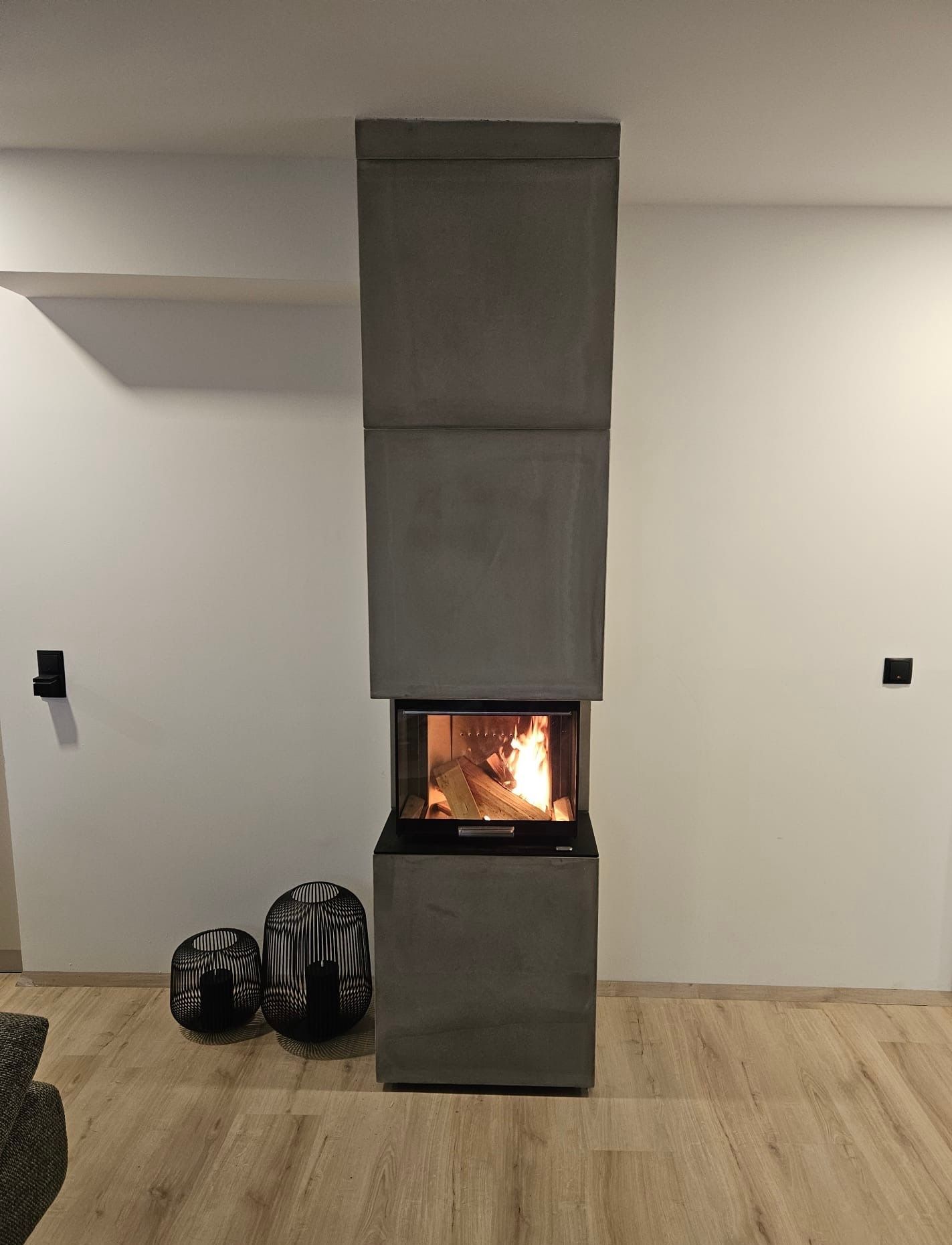 Modernes Zimmer mit einem hohen, grauen Kamin, der vor einer weißen Wand mit Holzboden brennt.