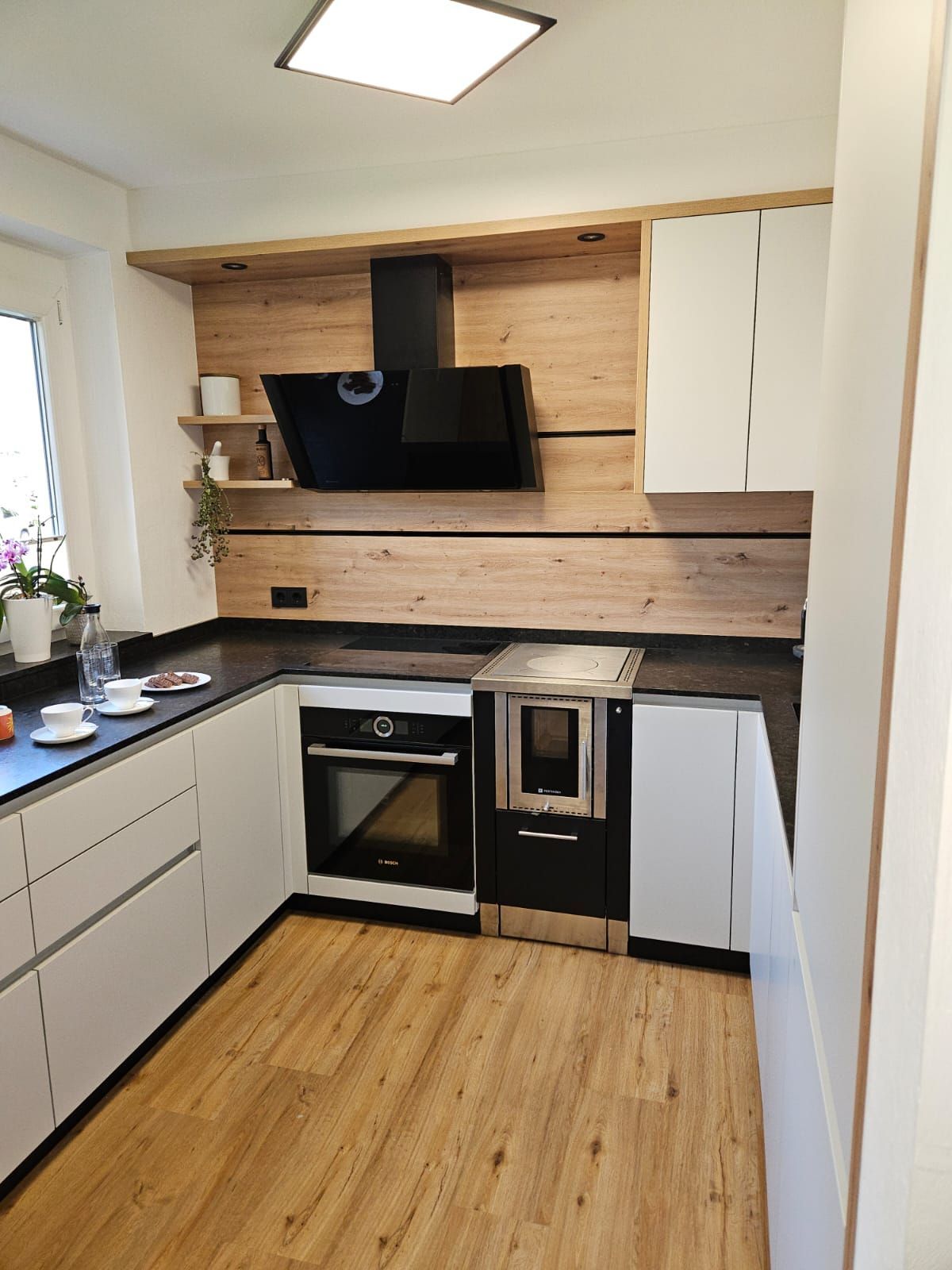 Moderne Küche mit hellen Holzschränken, schwarzen Arbeitsplatten, eingebautem Backofen und an der Wand montiertem Fernseher.