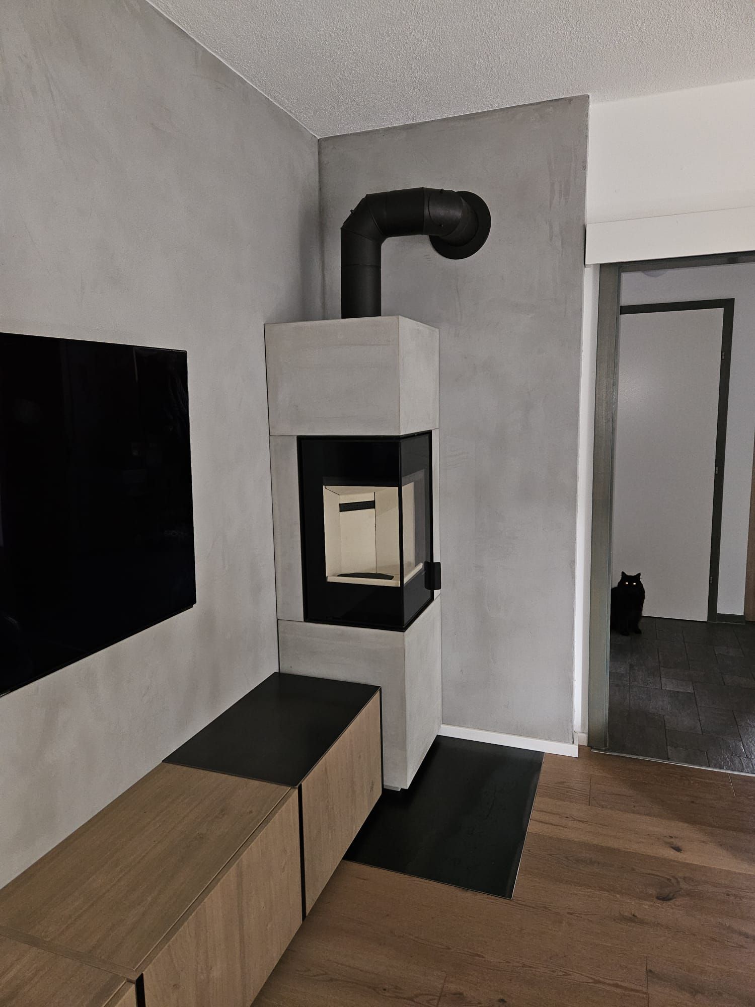 Modernes Wohnzimmer mit an der Wand montiertem Fernseher, Holzbank und einem weißen Holzofen mit schwarzem Rauchrohr.