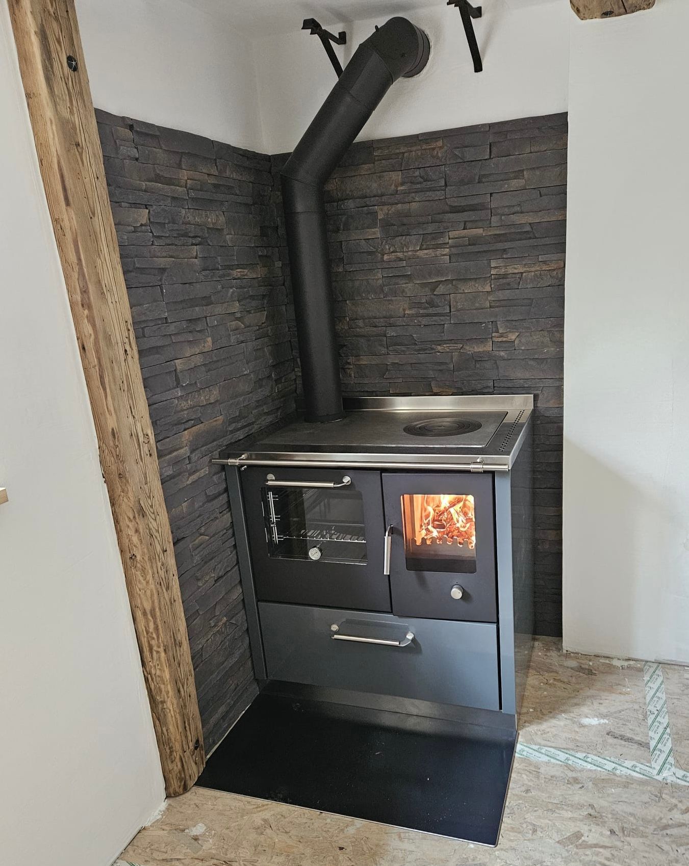 Holzofen mit schwarzem Rauchrohr in einer kleinen Ecknische mit dunklen Steinwänden.