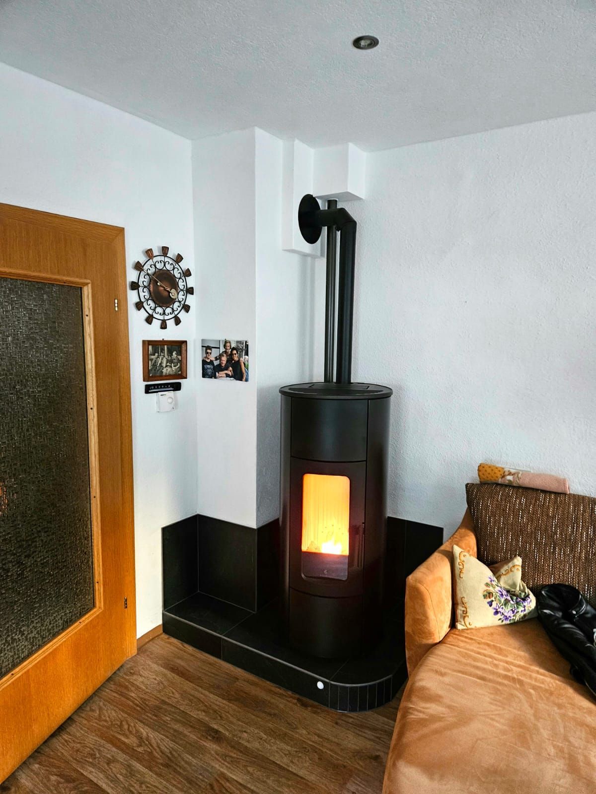 Gemütliches Zimmer mit schwarzem Holzofen, brennendem Kaminfeuer, hellbraunem Sofa und Holzboden