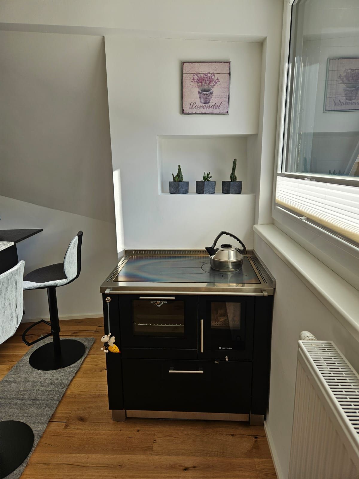 Kleine Büroküche mit Spüle, schwarzem Schrank, Regalen, Stuhl, Heizkörper und Jalousien.