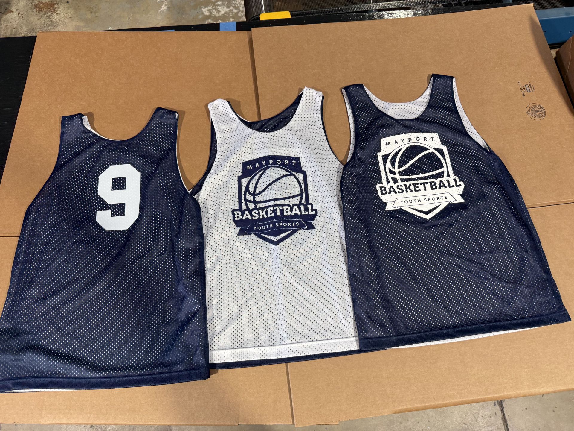Three navy and white mesh basketball jerseys laid on cardboard, one with number 9 and two with logos
