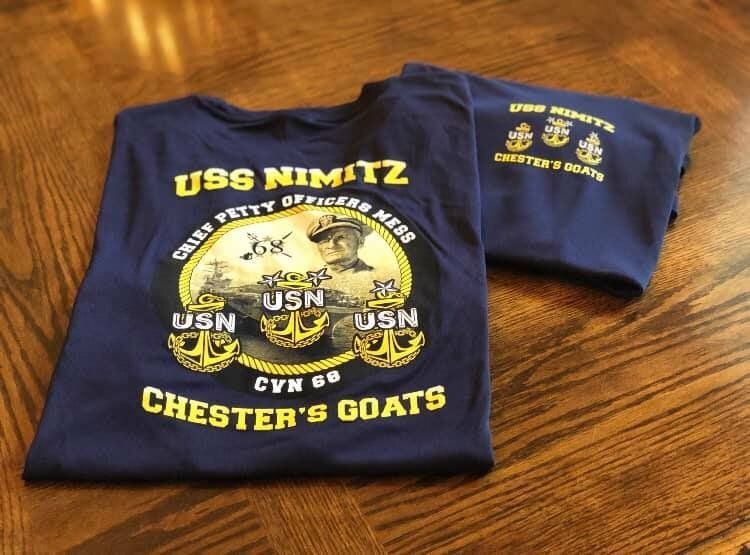 Two folded navy blue T-shirts with yellow military-style graphics on a wooden table