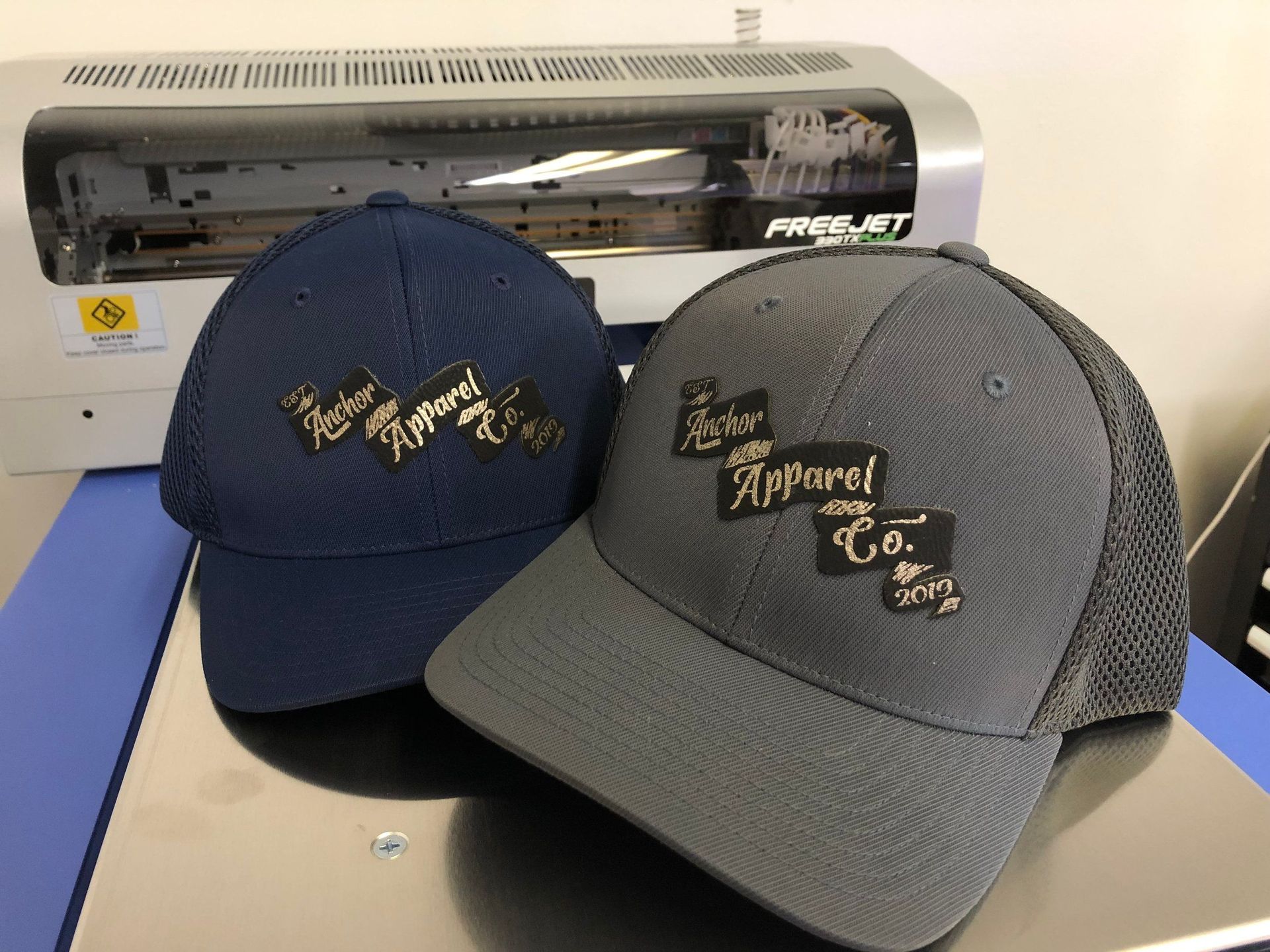 Two baseball caps on a table in front of a silver cutting machine, navy and gray with gold lettering