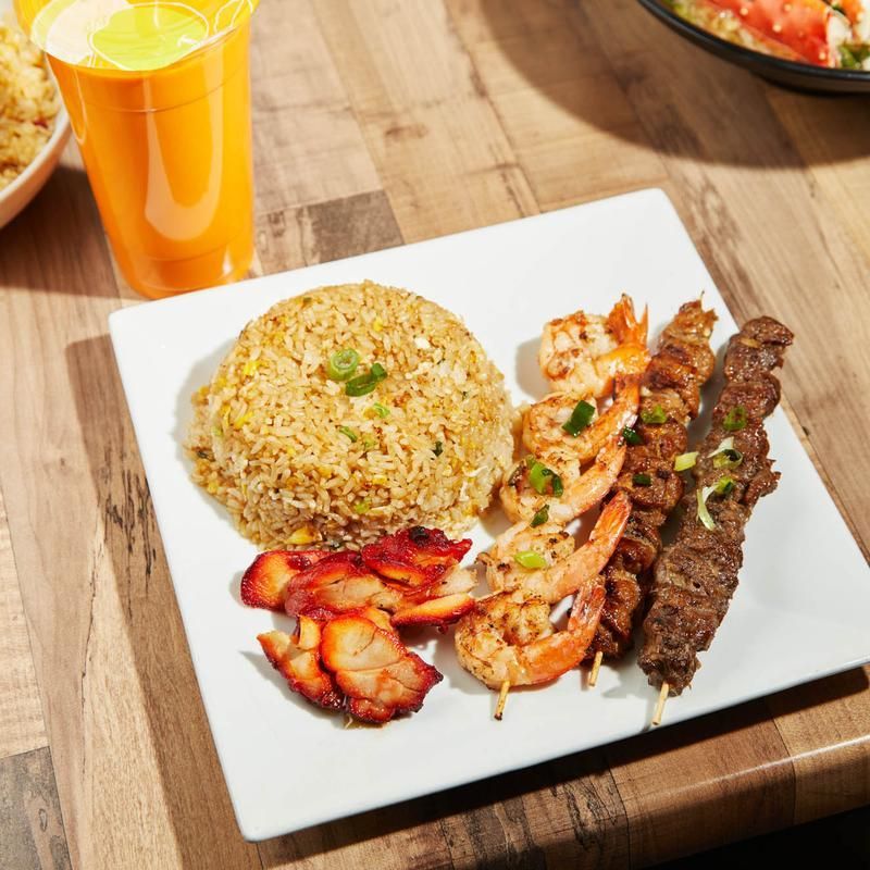 Plate of fried rice, grilled shrimp, skewered meat, and sliced chicken with a glass of orange drink on a table