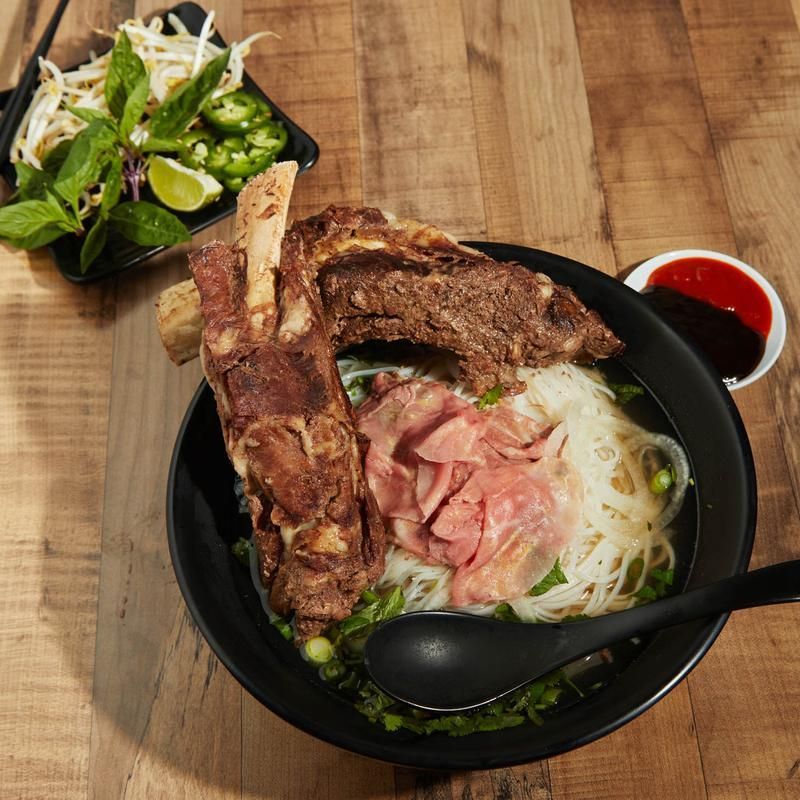 Ramen bowl with pork bones, sliced meat, noodles, greens, and a side salad on a wooden table