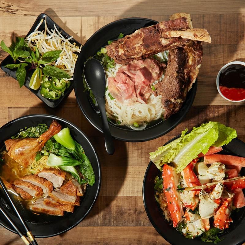 Four plated Asian-style noodle and meat dishes on a wooden table with chopsticks and a small sauce cup