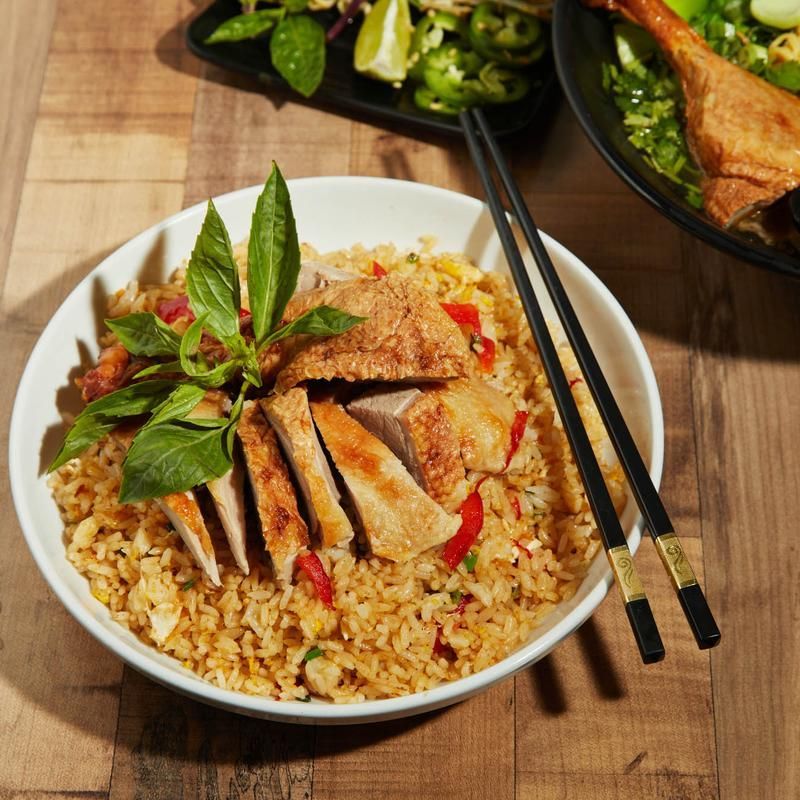 Bowl of sliced roasted meat over rice with basil, chopsticks, and side dishes on a wooden table
