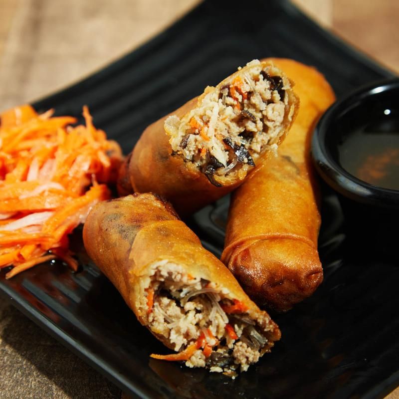 Crispy spring rolls on a black plate with a side of dipping sauce and shredded carrot salad