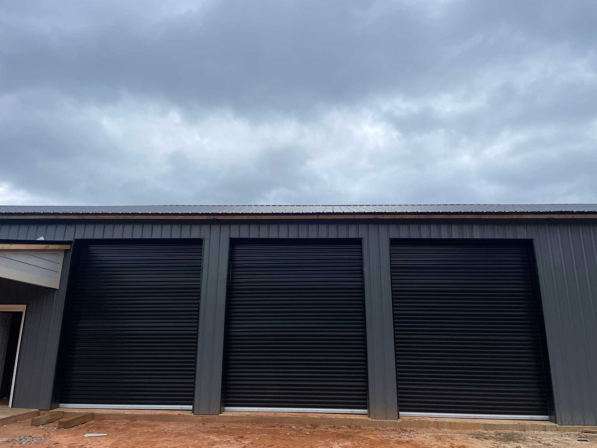 Three black roller doors on a grey building under a cloudy sky.