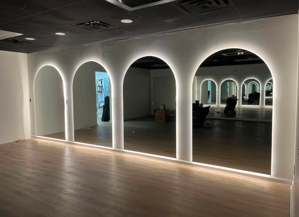 Modern room with glowing arched mirrors, wood floor, and dark ceiling; a person stands in the background.