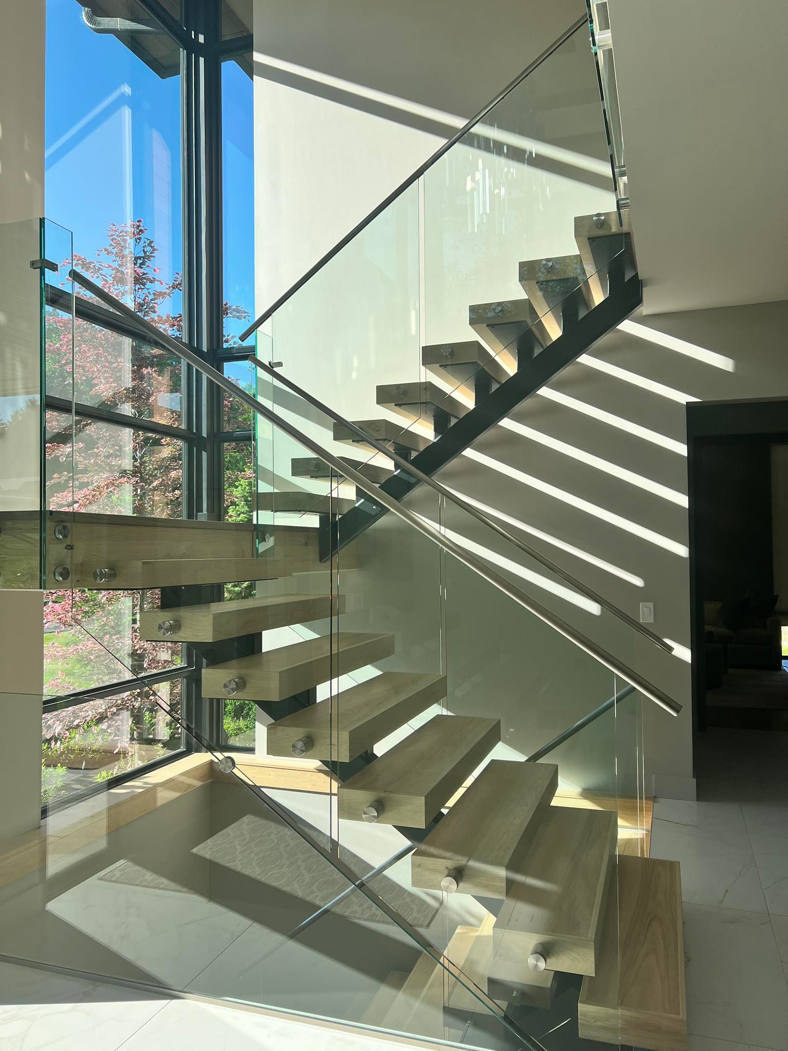 Modern glass staircase in a bright home, with sunlight casting striped shadows on the wall.