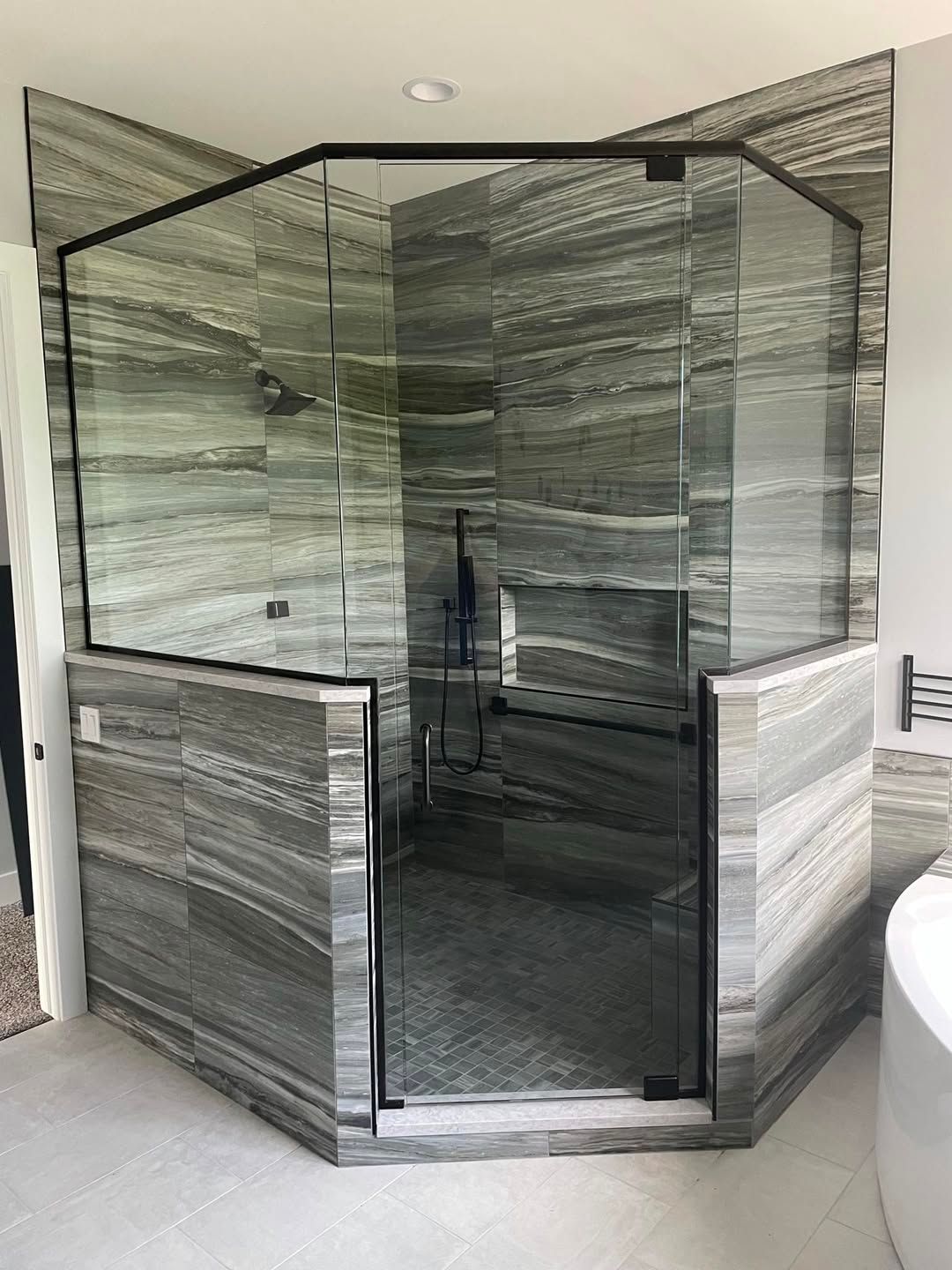 Corner glass shower enclosure with gray stone-pattern tile walls and a white tub nearby