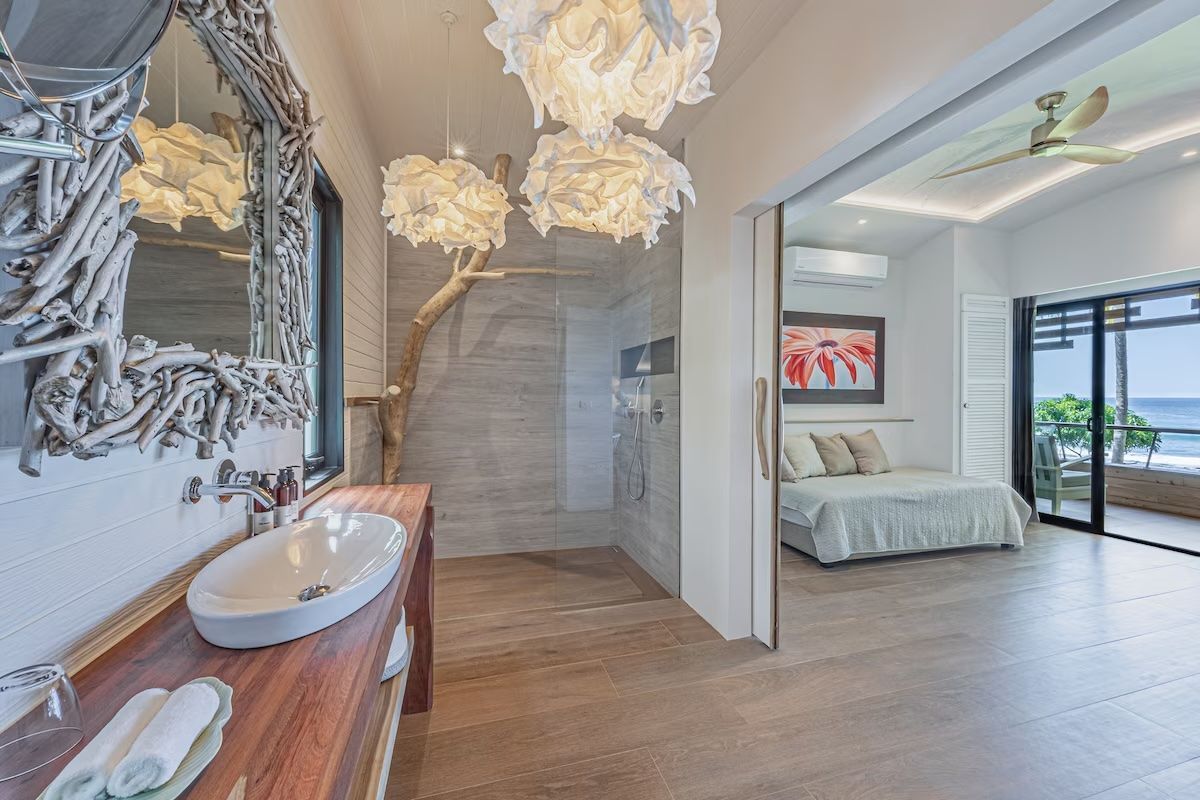 Salle de bains avec meuble-lavabo, ouverte sur une chambre avec vue sur l'océan. Comprend une douche à effet pluie, un cadre de miroir en bois flotté et un éclairage moderne.