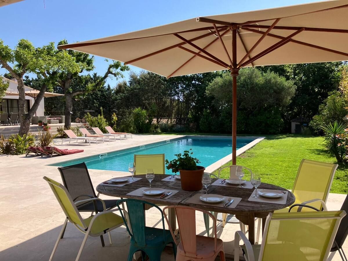 Table à manger extérieure dressée pour un repas, donnant sur une piscine et une pelouse verdoyante sous un grand parasol.