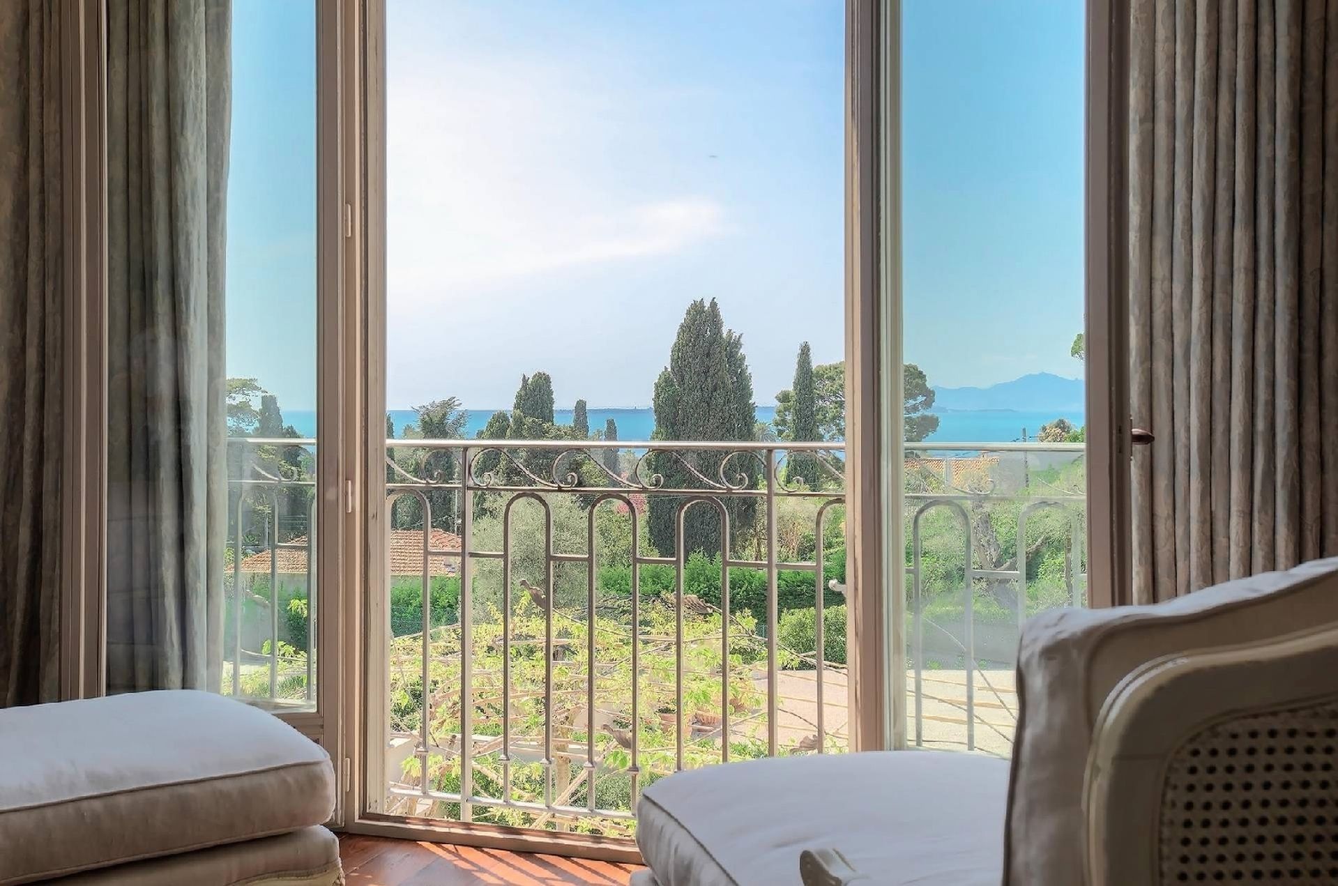 Vue depuis une chambre donnant sur un balcon avec des arbres, de la verdure et un plan d'eau au loin sous un ciel lumineux.