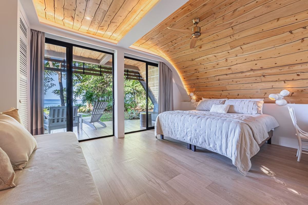 Chambre avec murs et plafond lambrissés, grandes fenêtres, lit avec linge de lit blanc et parquet clair.