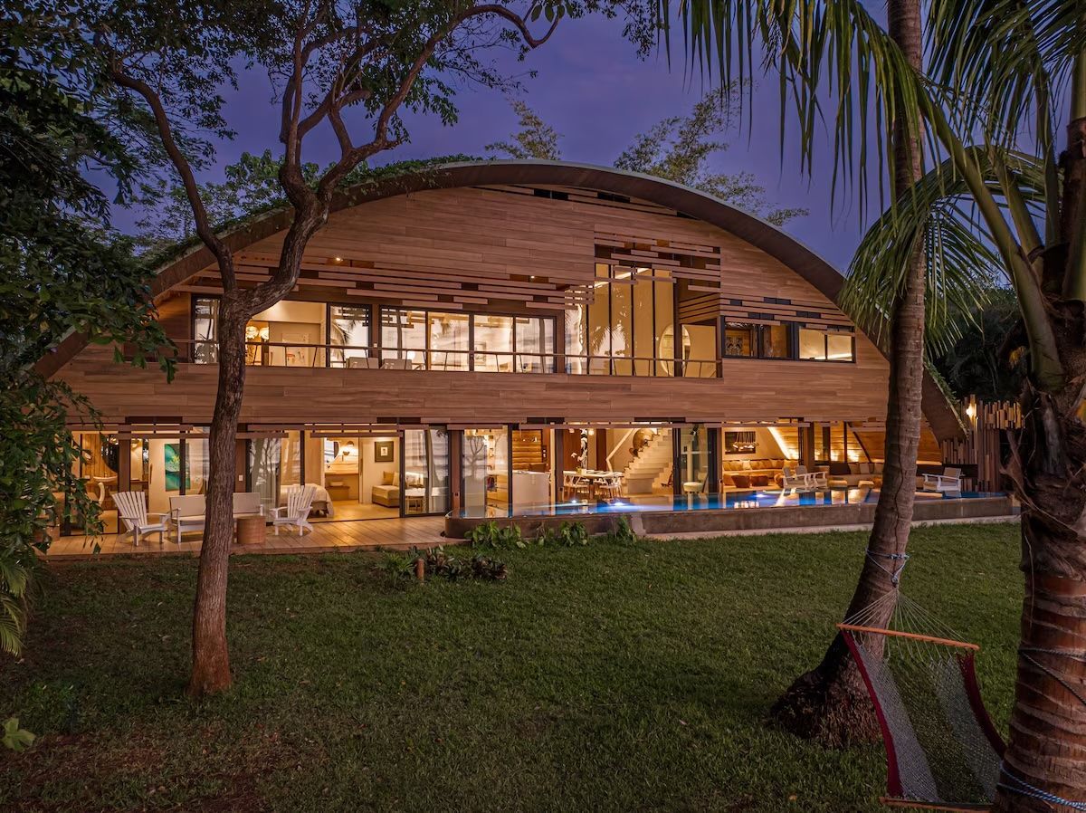 Maison en bois avec toit incurvé et grandes fenêtres, éclairée la nuit, avec piscine.