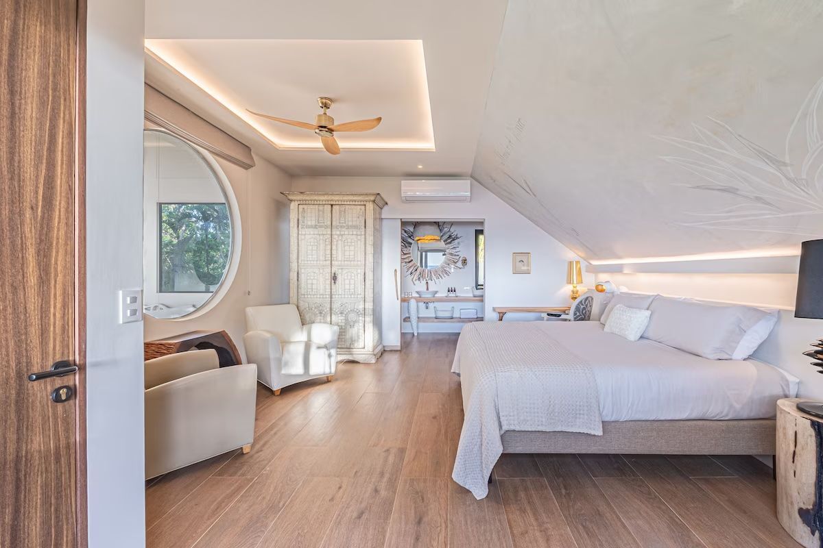 Chambre avec lit, placard, canapé, miroir et porte de salle de bain ; parquet clair, murs blancs.