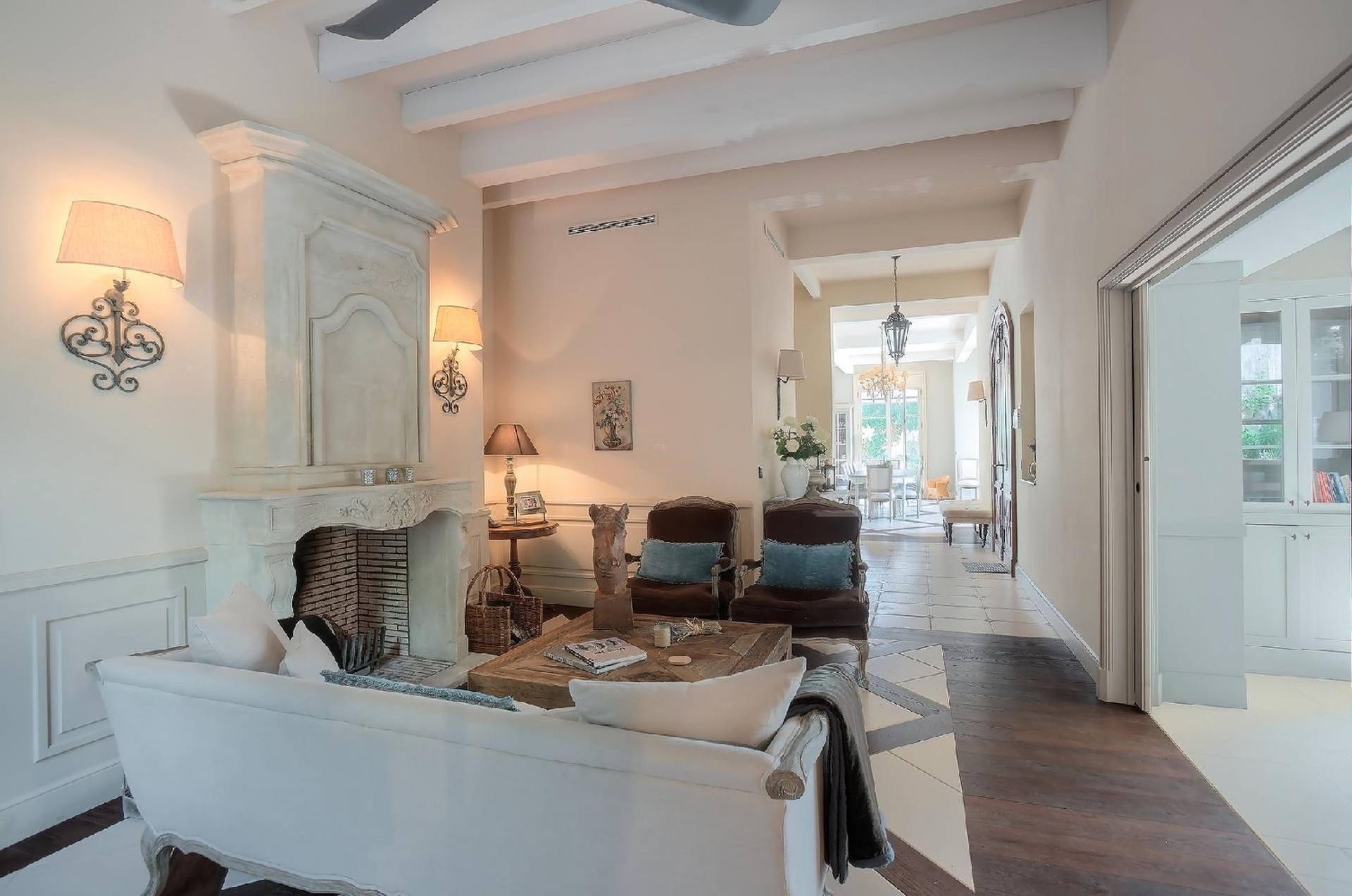 Salon avec cheminée blanche, poutres apparentes, coin salon et porte donnant sur une autre pièce.
