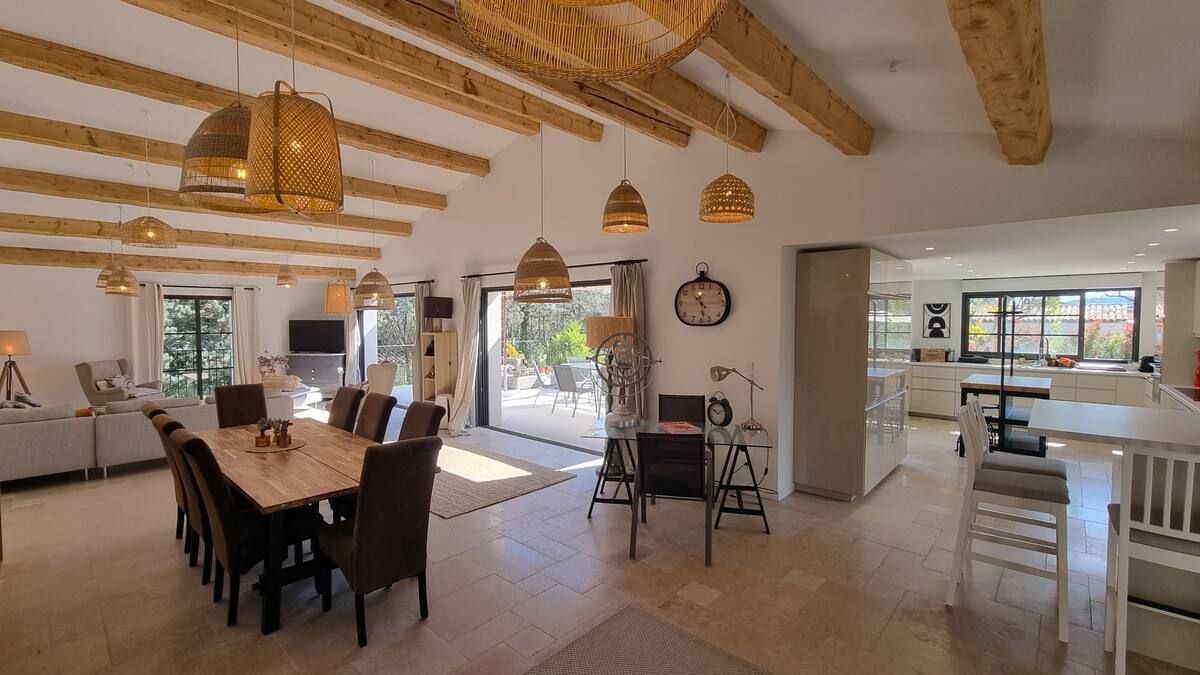 Vaste espace de vie ouvert avec table à manger, cuisine et portes-fenêtres donnant sur le patio. Poutres apparentes et suspensions lumineuses.