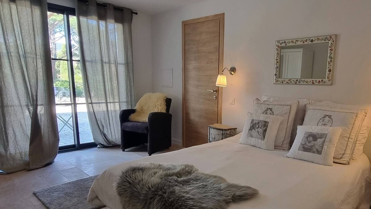 Chambre avec un lit, un fauteuil près de la fenêtre et un miroir mural. Linge de lit blanc, tons neutres.