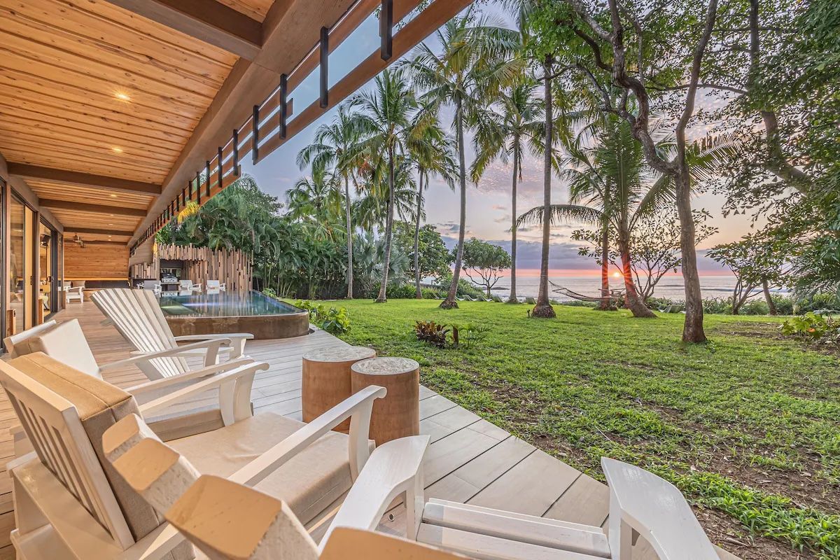 Terrasse en bois avec chaises blanches, vue sur la pelouse, les arbres et l'océan au crépuscule.