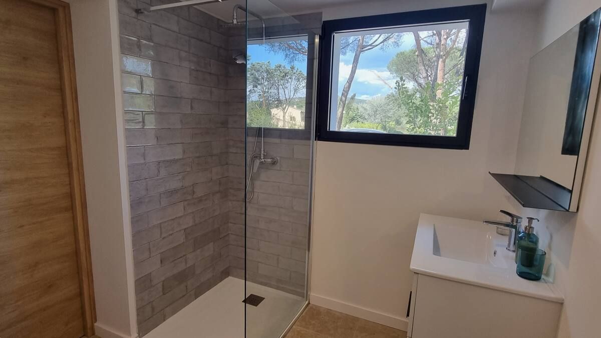 Salle de bains avec douche carrelée, meuble-lavabo et fenêtre donnant sur les arbres.