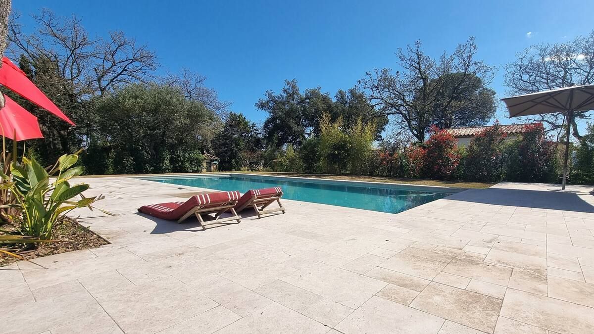 Piscine avec deux chaises longues, parasols rouges et arbres sous un ciel bleu azur.