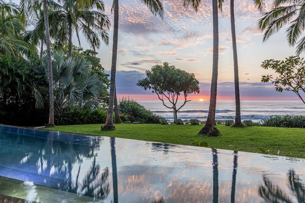 La piscine à débordement reflète le coucher de soleil sur l'océan, encadrée par des palmiers et de la pelouse verdoyante.
