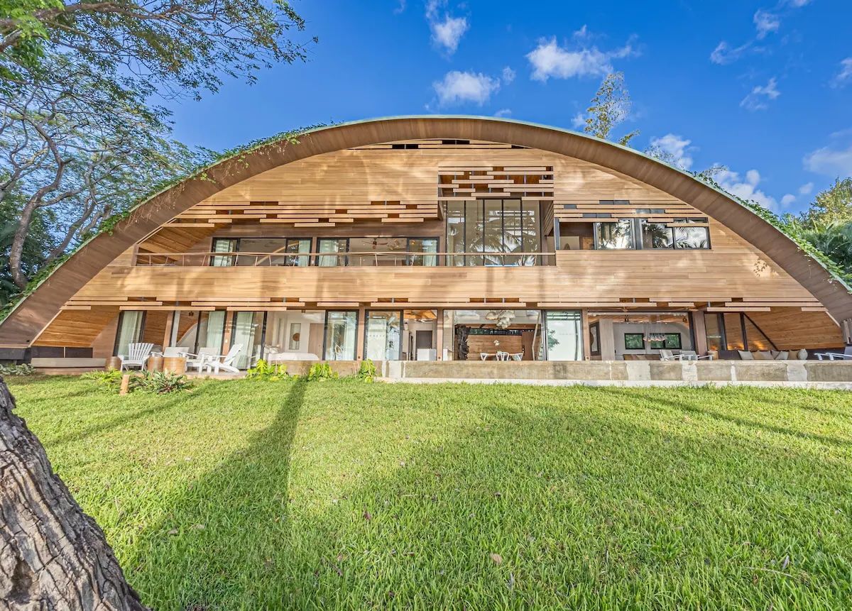 Maison en bois au toit voûté sur une pelouse verte ; murs de verre, ciel bleu.