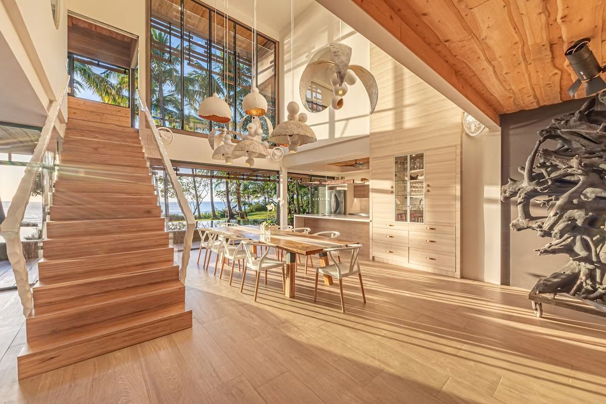 Un escalier en bois mène à une maison moderne et lumineuse comprenant un coin repas, une cuisine et une vue sur l'océan à travers de grandes fenêtres.