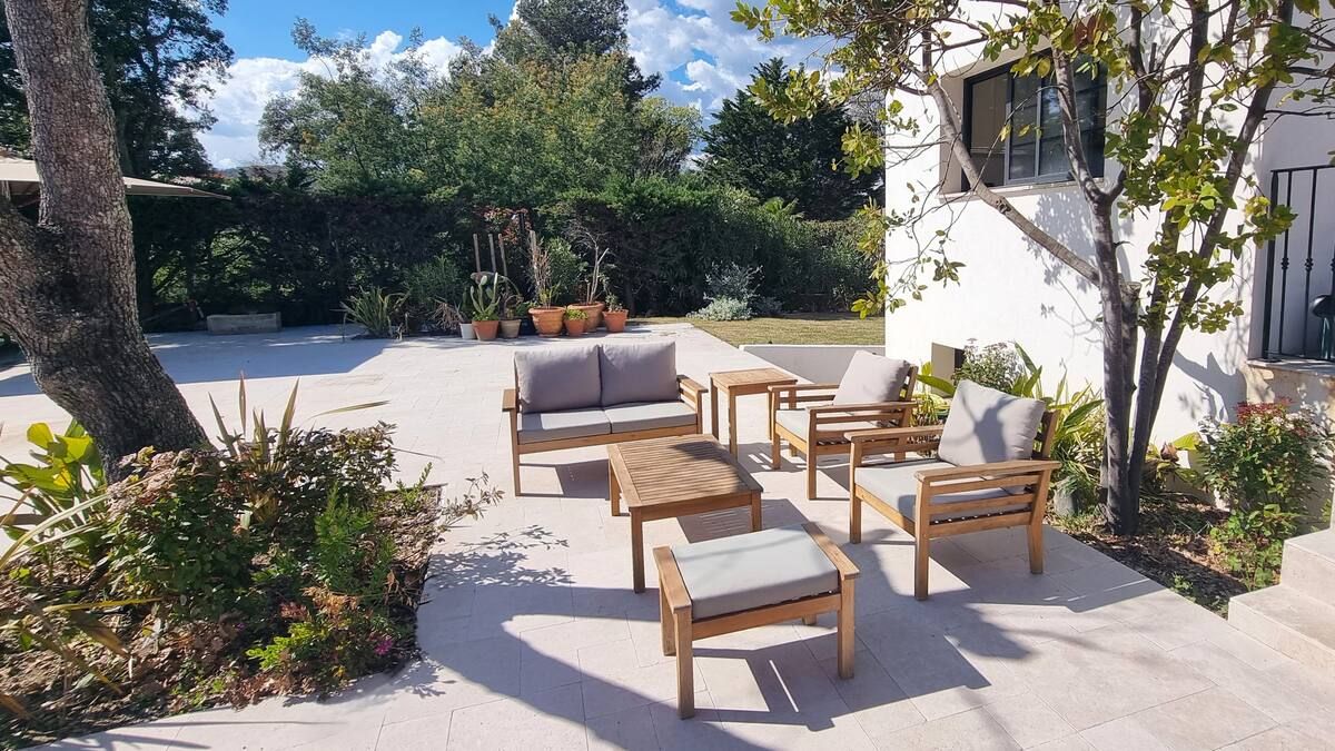 Coin salon extérieur avec mobilier en bois, coussins gris et un bâtiment blanc.