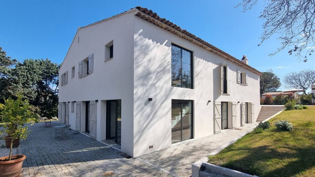 Maison blanche à deux étages avec encadrements de fenêtres foncés, terrasse en pierre, pelouse verte et ciel bleu.