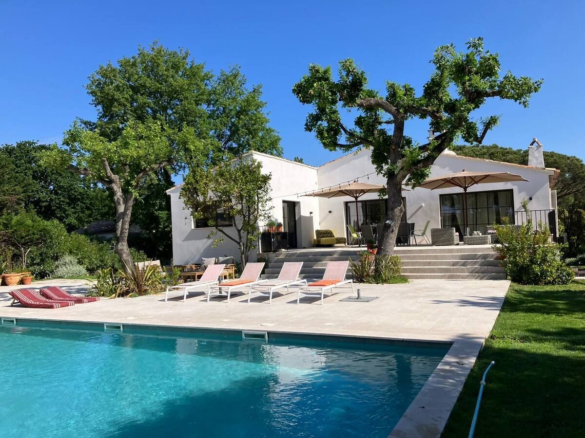 Villa blanche avec piscine, chaises longues et arbres sous un ciel bleu azur.