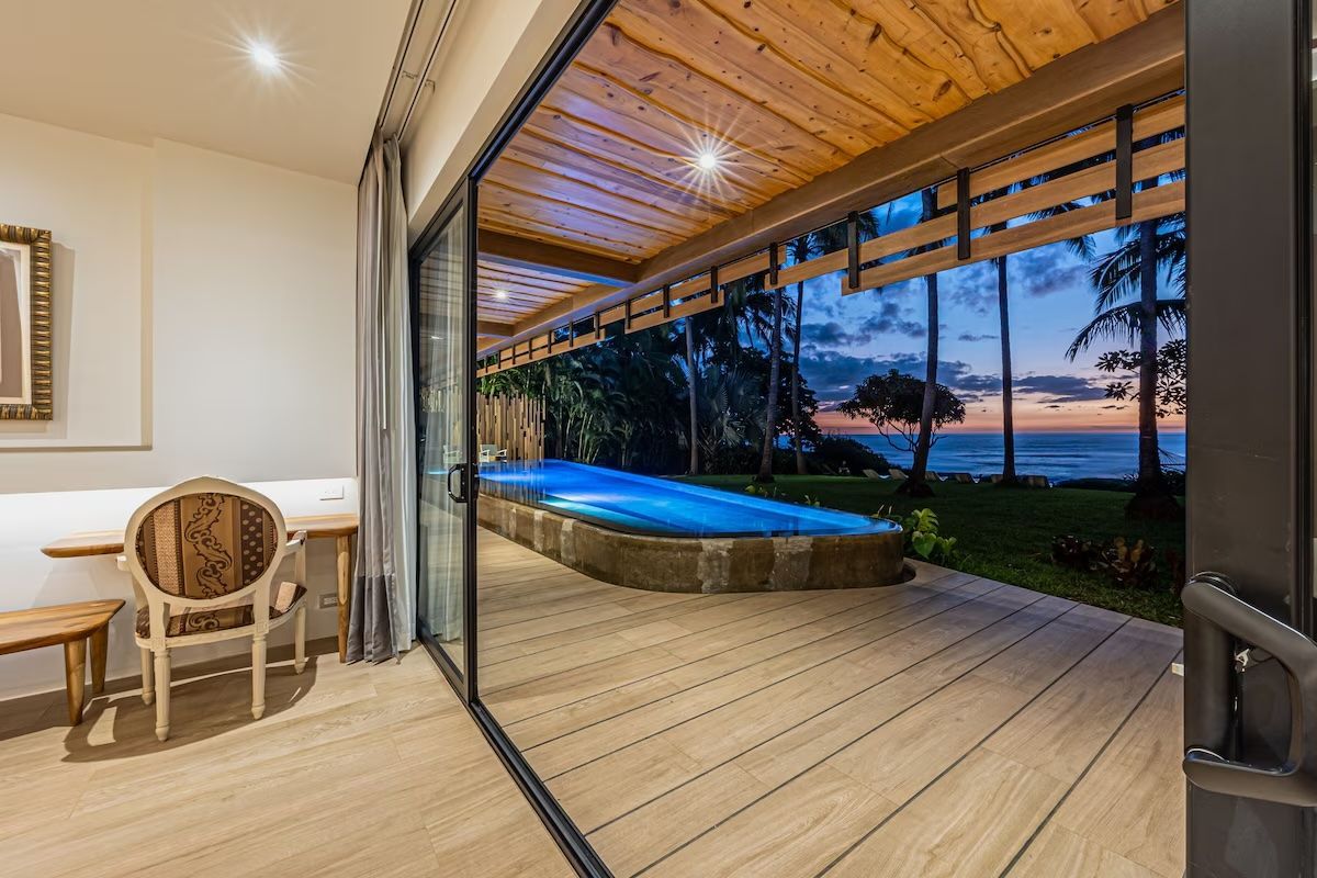Vue intérieure-extérieure sur une piscine et l'océan au coucher du soleil, à travers des portes coulissantes en verre. Terrasse et plafond en bois ; fauteuil à l'intérieur.