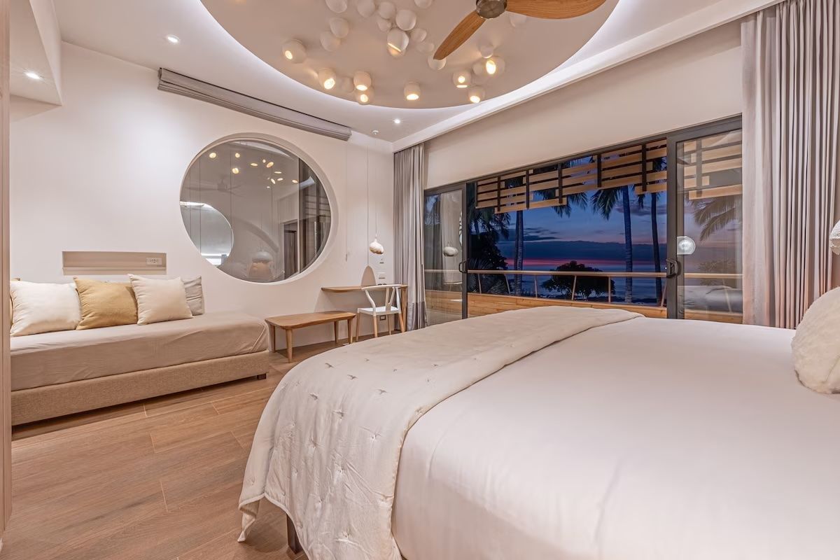 Chambre avec fenêtre ronde, grand lit et vue sur l'océan à travers une porte coulissante en verre.