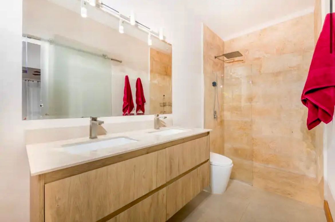 Salle de bains moderne avec double vasque, grand miroir et douche à l'italienne. Bois clair et pierre beige. Serviettes rouges.