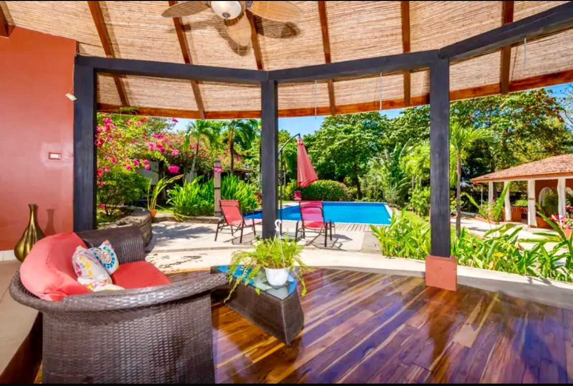 Terrasse extérieure avec sièges, donnant sur une piscine et un jardin luxuriant sous un toit de chaume.