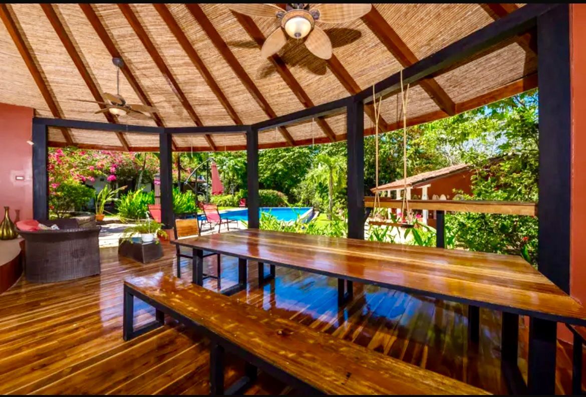Terrasse couverte avec une grande table en bois, des bancs et une piscine en arrière-plan.