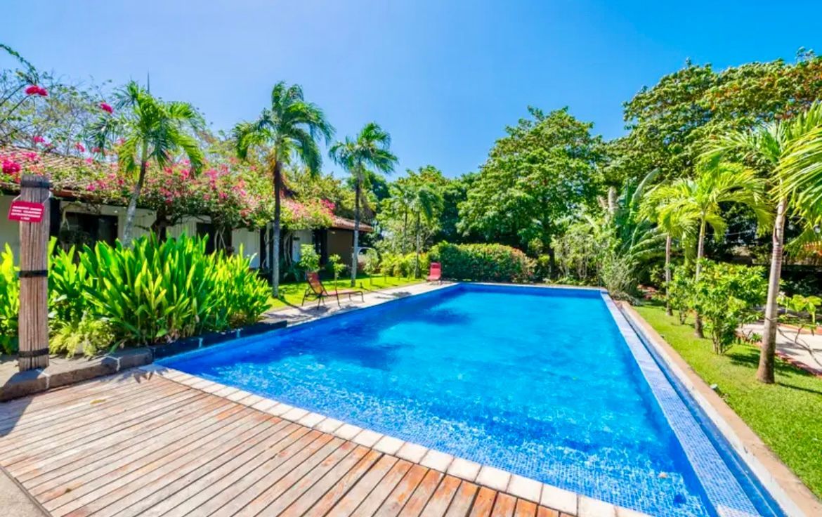 Piscine dans un cadre tropical : eau turquoise, pelouse verte, palmiers et bâtiments aux toits de tuiles rouges.