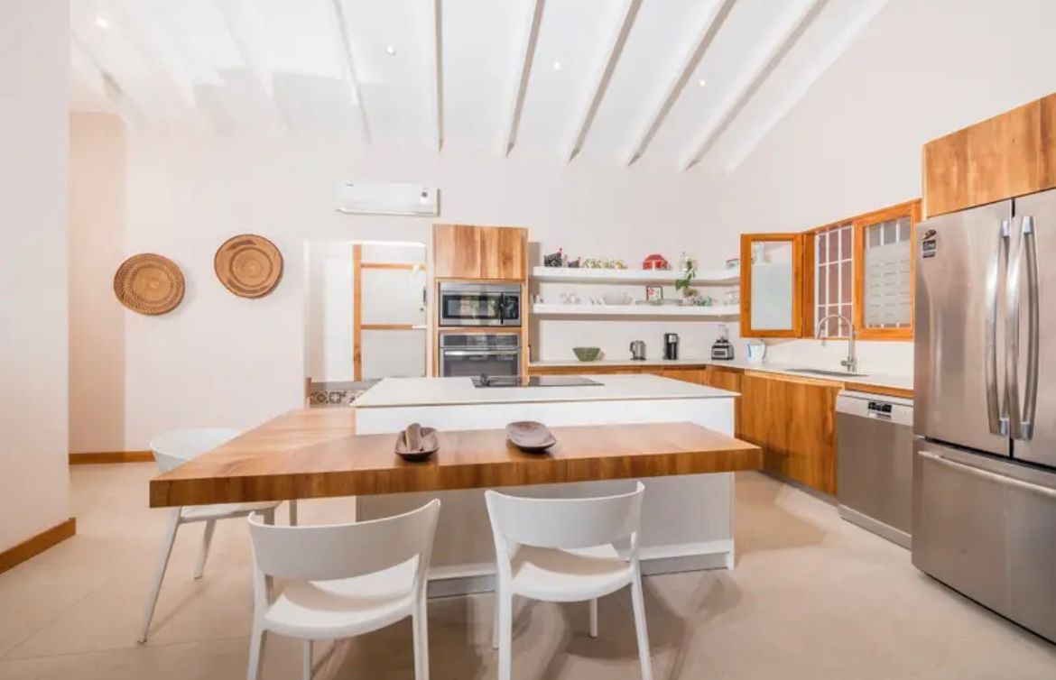 Cuisine moderne avec îlot en bois, chaises blanches, appareils électroménagers en acier inoxydable et armoires en bois.
