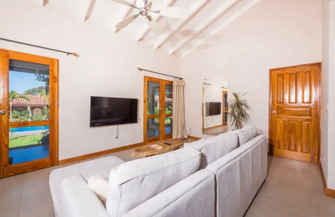 Salon avec canapé blanc, télévision, portes et plafond en bois, et vue sur une piscine.