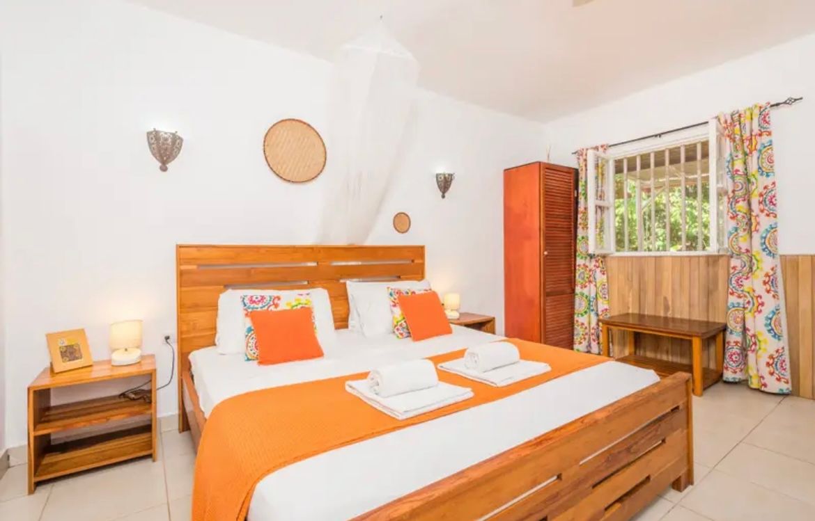 Chambre avec un lit en bois, des touches d'orange et une fenêtre ornée de rideaux à fleurs.