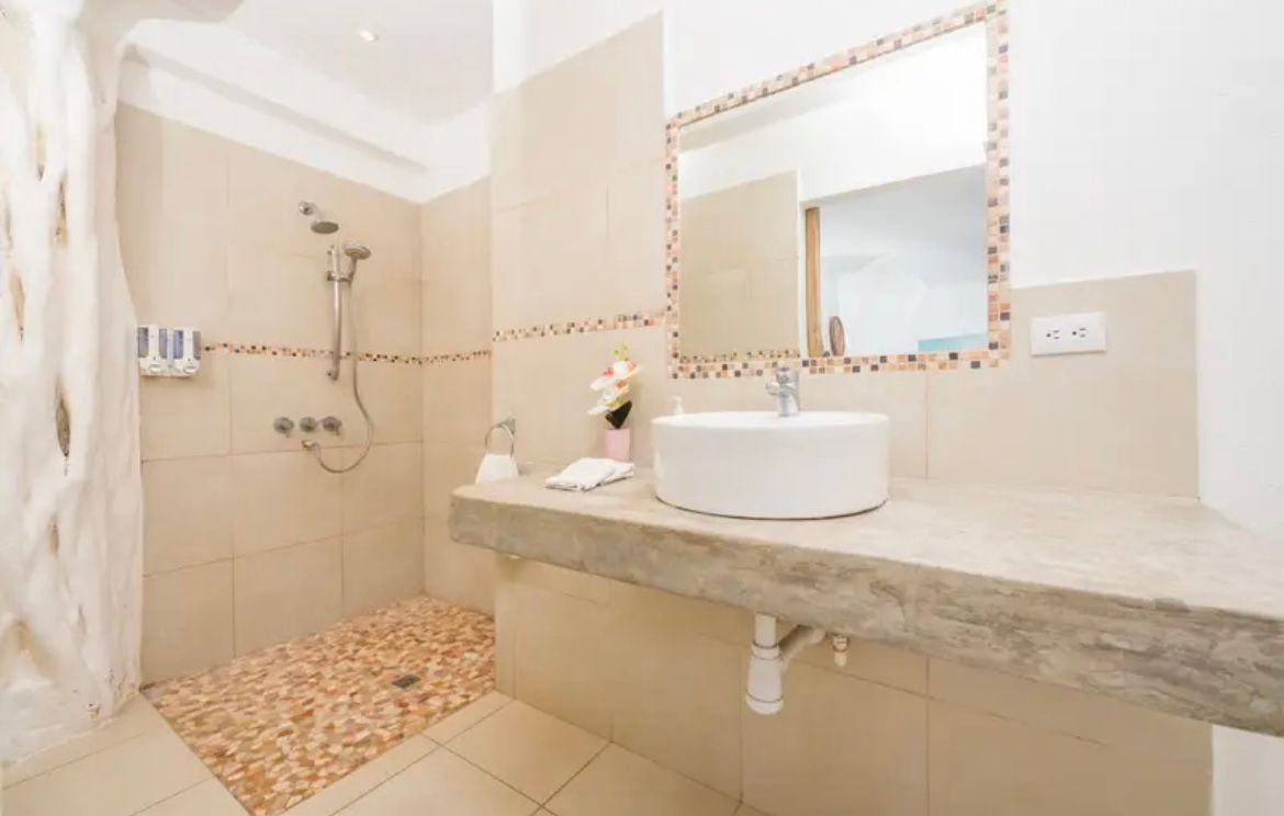 Salle de bains beige avec douche, sol en pierre, comptoir en béton, lavabo rond, miroir et articles de toilette.